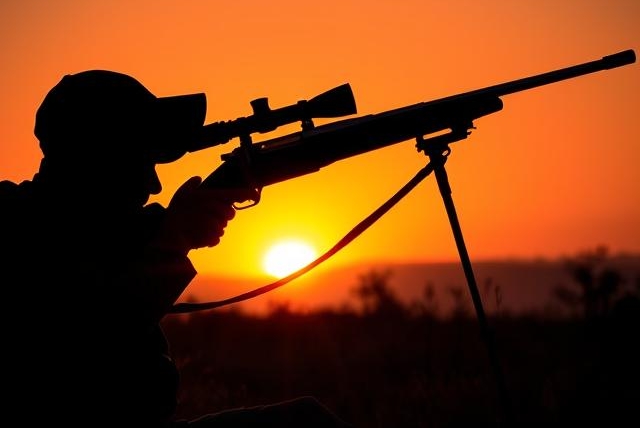 South African hunter silhouetted against sunrise, aiming rifle on tripod in the veld